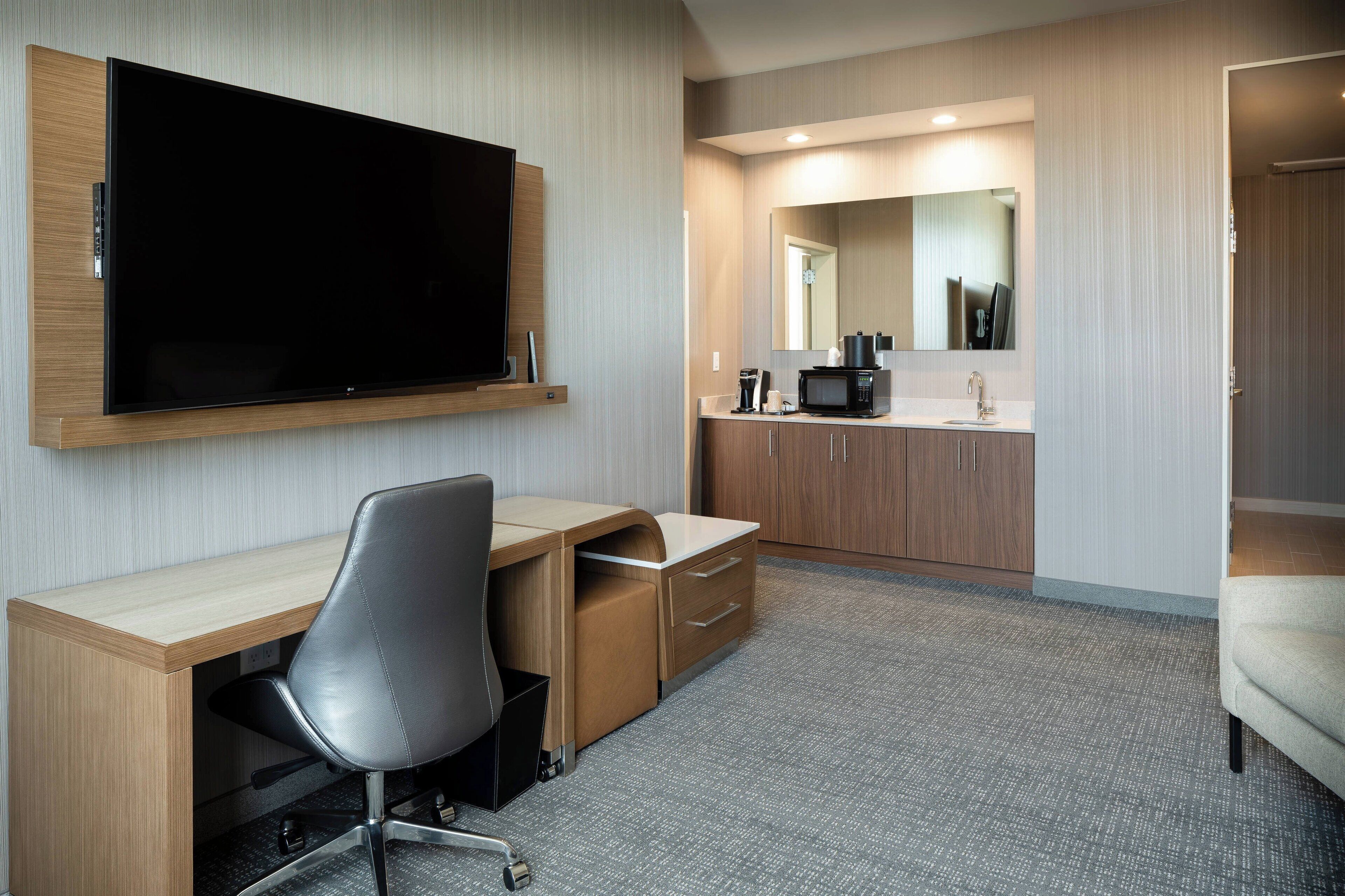 Habitación de hotel con televisión de pantalla plana montada en la pared, un escritorio con silla, microondas y un lavabo con espejo arriba.