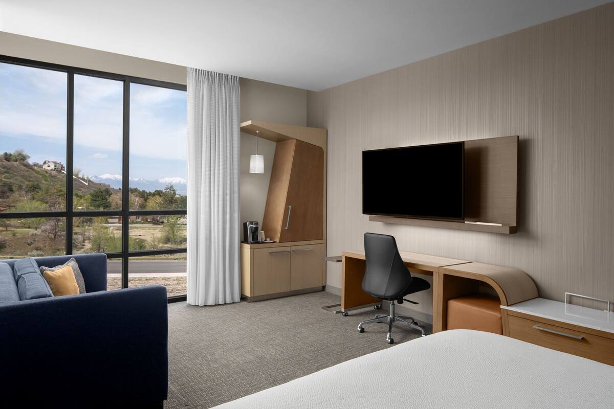 A hotel room with a large window, a bed, a desk, a chair, and a TV mounted on the wall.