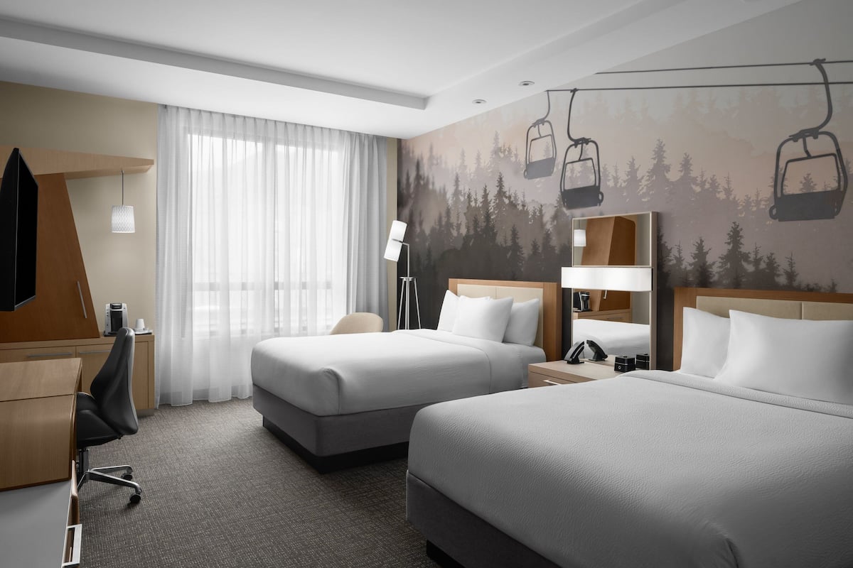 A hotel room with two beds, a desk, a chair, a lamp, and a ski lift mural on the wall.