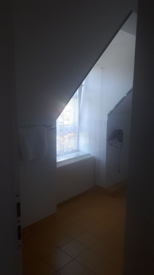 A dimly lit attic room with a sloped ceiling, a radiator, and a window with lace curtains. - Hotel Pension Am Engelsberg (Sommerach)