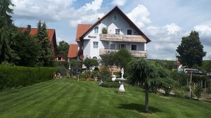 A two-story hotel with a red roof, a well-maintained lawn, and a statue in the front yard. - Hotel Pension Am Engelsberg (Sommerach)
