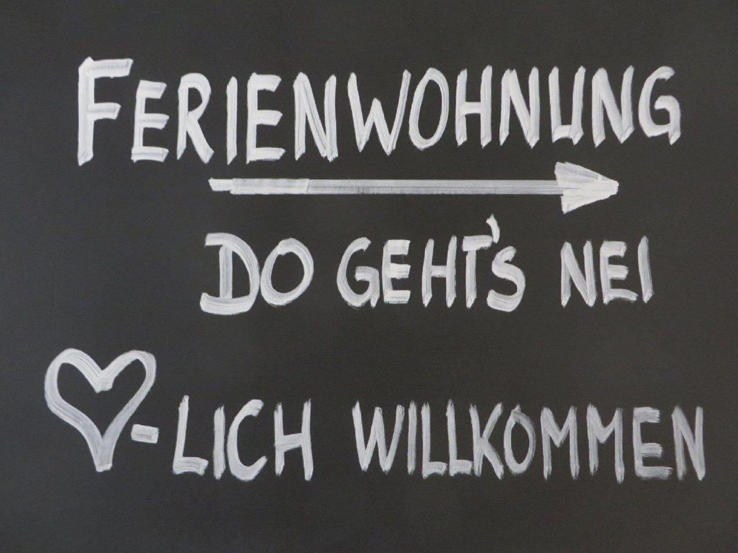 Um letreiro com o texto 'Feriendorfwohnung DO GEHT'S NEI LICH WILLKOMMEN' e um símbolo de coração.