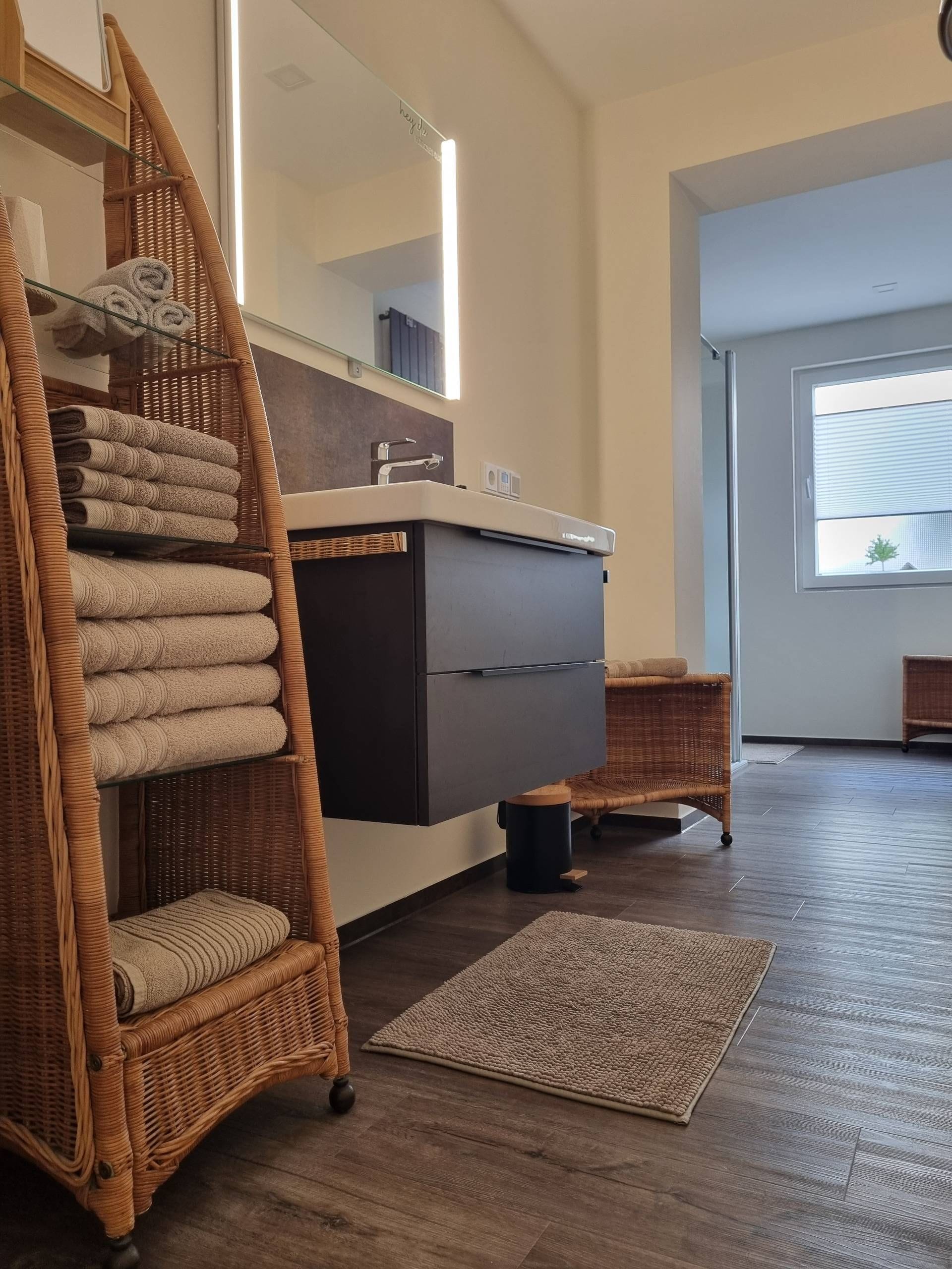 A modern bathroom with a large mirror, a dark-colored vanity, and a wicker chair with towels.