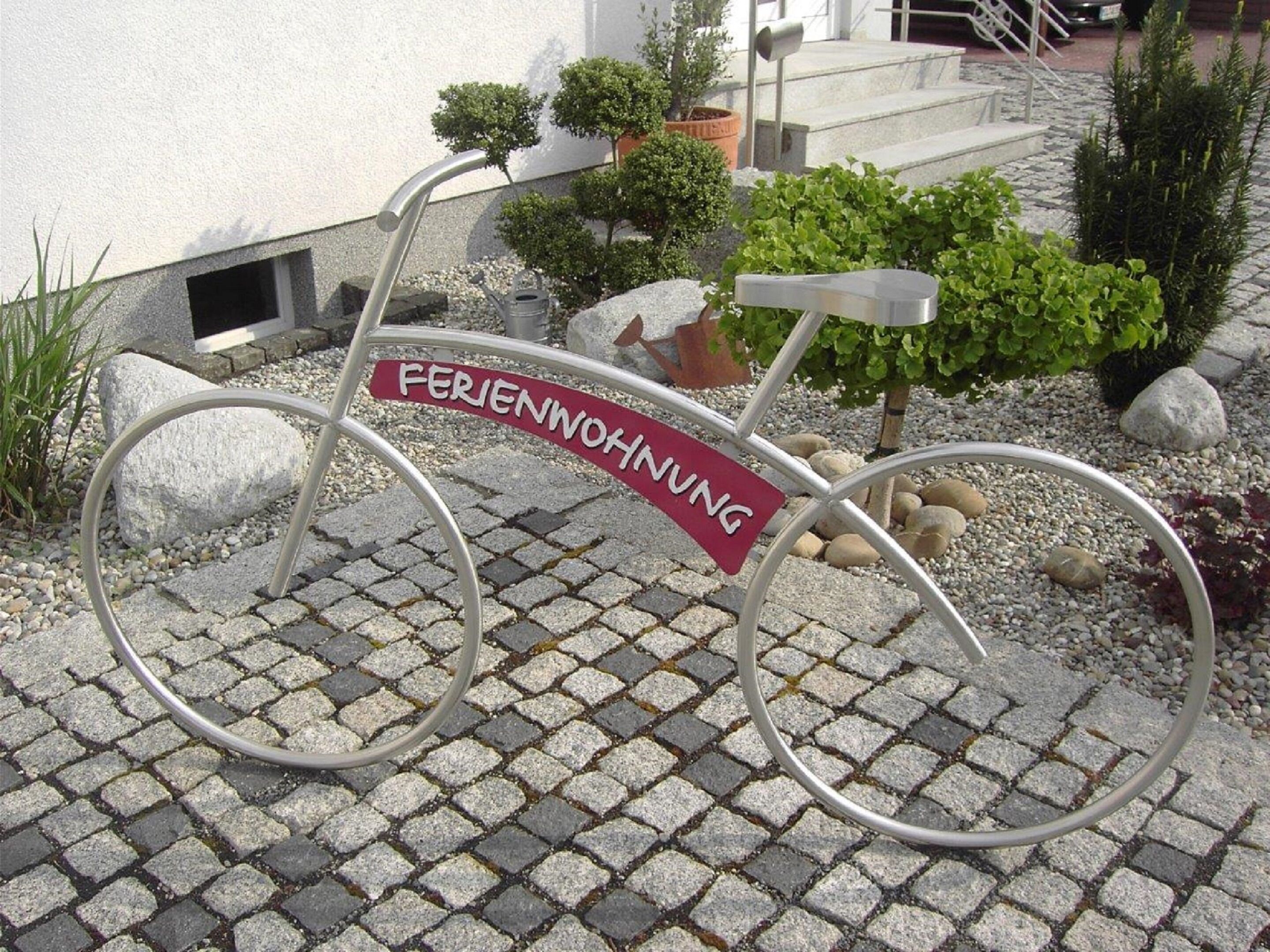Una bicicleta con un cartel que dice "FERIENWOHNUNG" frente a un edificio con entrada empedrada y plantas en macetas.