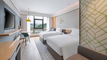 A hotel room with a large bed, a desk, a chair, a television, and a window with a view of buildings and greenery.
