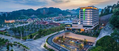 Un hotel moderno con un llamativo letrero verde, un estacionamiento y una vista panorámica de un parque de atracciones cercano.