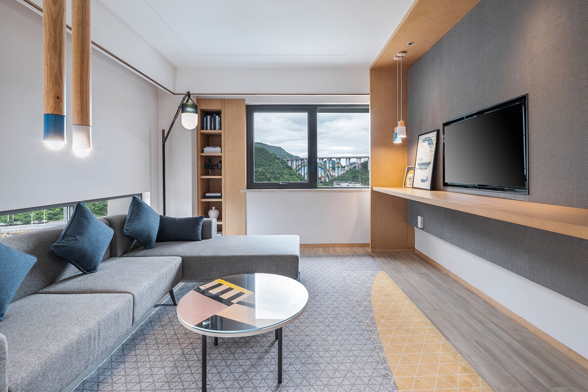A modern living room with a sofa, a glass coffee table, a bookshelf, a TV mounted on the wall, and a view of a bridge through the window.