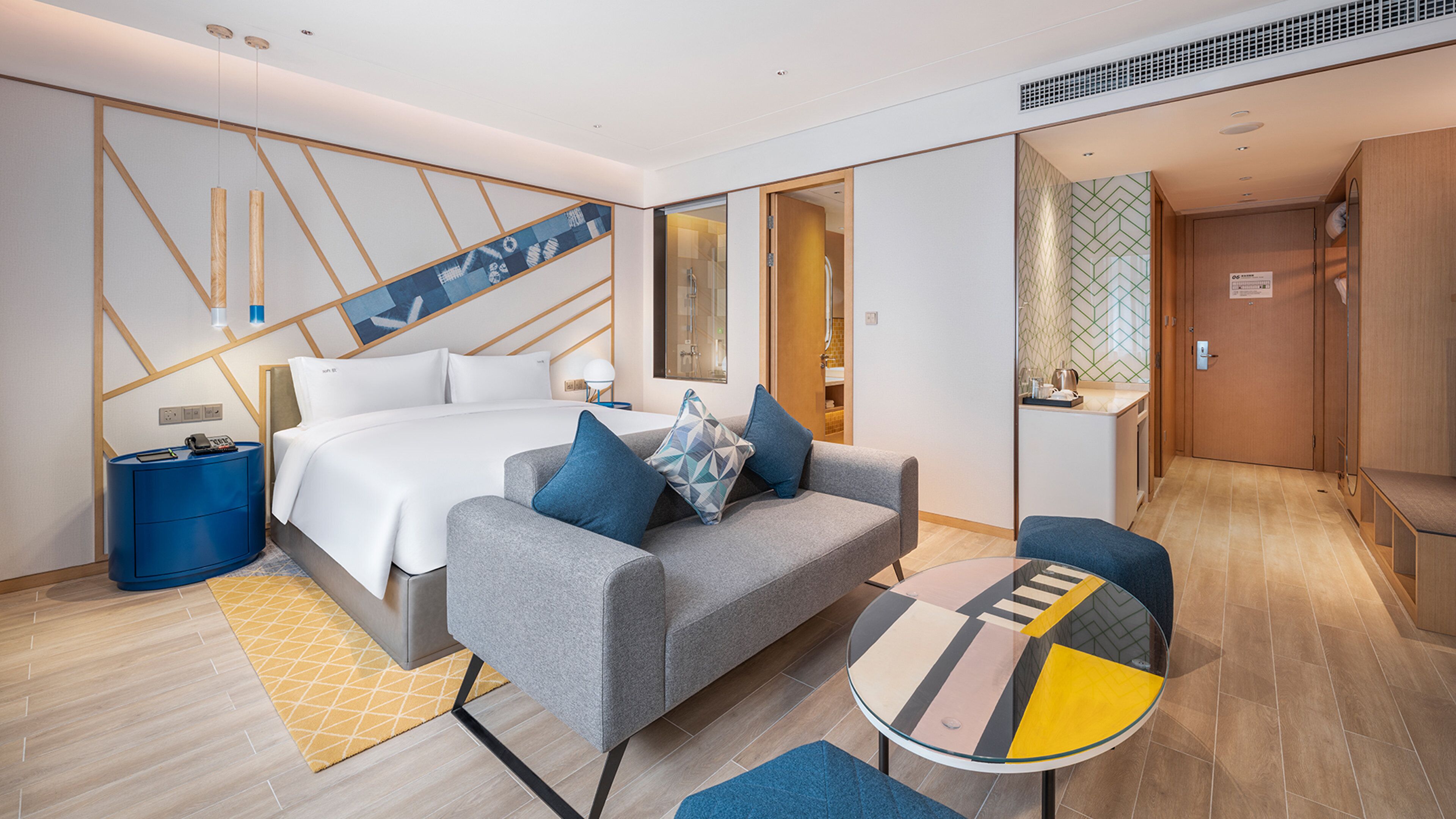 A modern hotel room with a bed, a sofa, a small table, and a bathroom visible through an open door.
