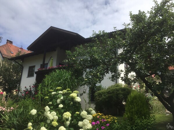 A house with a sloped roof, a balcony with flowers, and a garden with various plants and flowers.