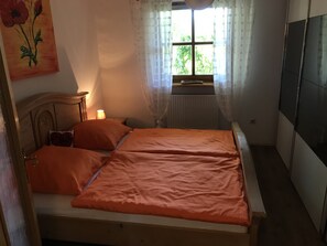 A bedroom with a wooden bed, orange bedding, a bedside lamp, a window with sheer curtains, and a painting on the wall.