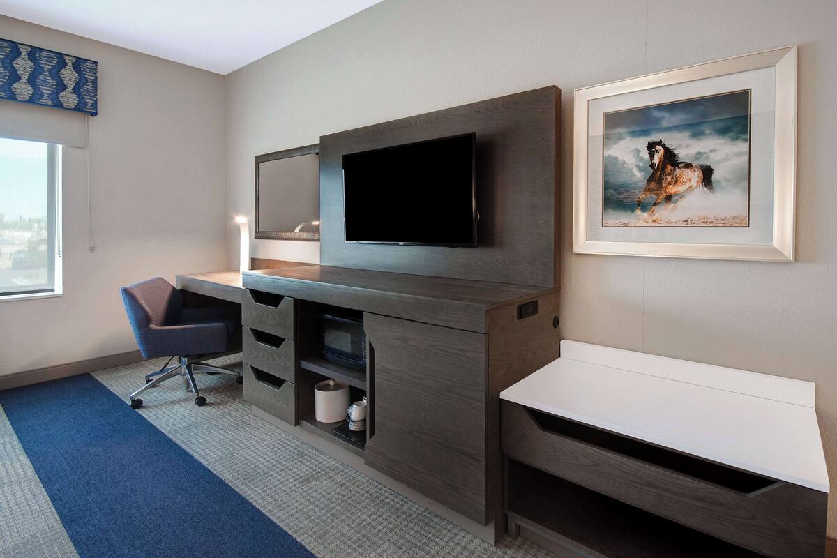 A modern hotel room with a large flat-screen TV mounted on the wall, a desk with a chair, a white bench, and a framed picture of a horse.