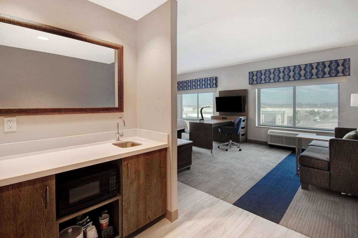 A modern hotel room with a kitchen area, a large window with a city view, a flat-screen TV, and a comfortable seating area.
