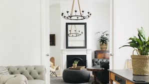 A living room with a grey sofa, a wooden dining table with stools, a black leather armchair, a black ottoman, a fireplace, a chandelier, and a large mirror. - Castlebar - Superior Boutique Accomodation - Steps to Pakington Street (Geelong West)