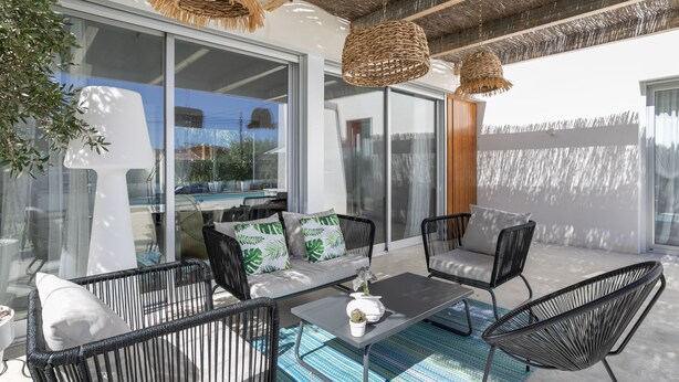 An outdoor patio with wicker furniture, a glass door leading to a pool, and a view of the surrounding area.