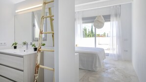 A modern bedroom with a large window, a bed, and a bathroom with a sink and plants.