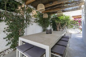 Un espace de restauration en plein air avec une table blanche et des bancs gris dotés de coussins, entouré de verdure et d’un mur blanc.