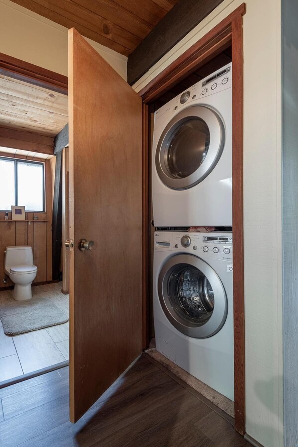 Une buanderie équipée de deux machines à laver à chargement frontal superposées, d’une porte en bois et de toilettes visibles au fond.