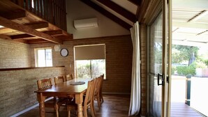 Un comedor con una mesa y sillas de madera, pared de ladrillo, ventana con persianas y puerta corredera de vidrio que da a una terraza exterior.