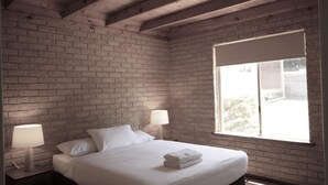 Un dormitorio con pared de ladrillo, un ventanal grande, una cama con ropa blanca y dos lámparas de noche.