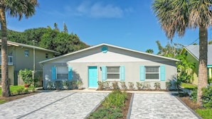 A single-story building with a flat roof, light-colored exterior, and blue shutters. There is a paved driveway leading to the entrance, and a small garden area with various plants.