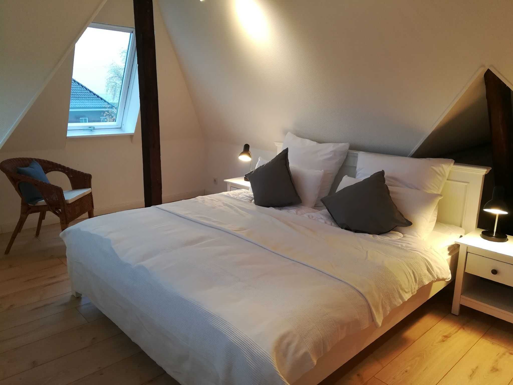 Une chambre avec un plafond en pente, un lit avec une literie blanche et des oreillers foncés, une chaise, une table de chevet avec une lampe et une fenêtre laissant entrer la lumière naturelle.