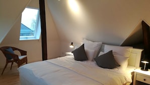 Ein Schlafzimmer mit schrägen Wänden, einem Bett mit weißer Bettwäsche und dunklen Kissen, einem Sessel, einem Nachttisch mit Lampe sowie einem Fenster, das für natürliches Licht sorgt.