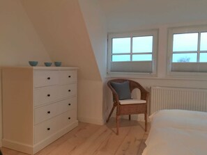 Un dormitorio con un tocador blanco, una silla de mimbre, una cama y dos ventanas con persianas.