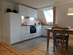 Una cocina moderna con armarios blancos, horno integrado y una zona de comedor con mesa y silla de madera.