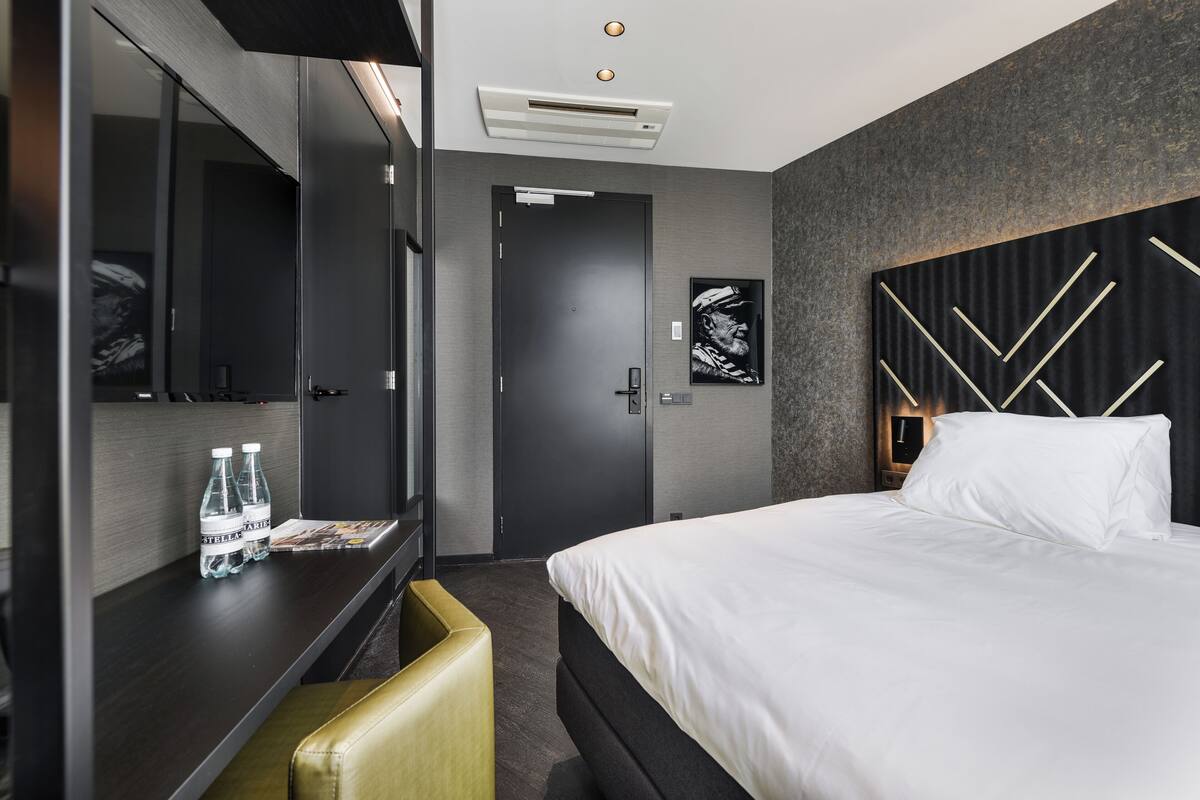 Ein modernes Hotelzimmer mit einem großen Bett, einem Schreibtisch mit Stuhl, einem Kleiderschrank und einem Spiegel. Auf dem Schreibtisch stehen Wasserflaschen und an der Wand hängt ein gerahmtes Bild.