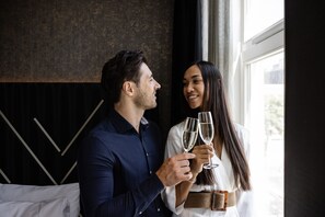 A couple holding champagne glasses in a room with a modern interior, a window, and a bed with white bedding.