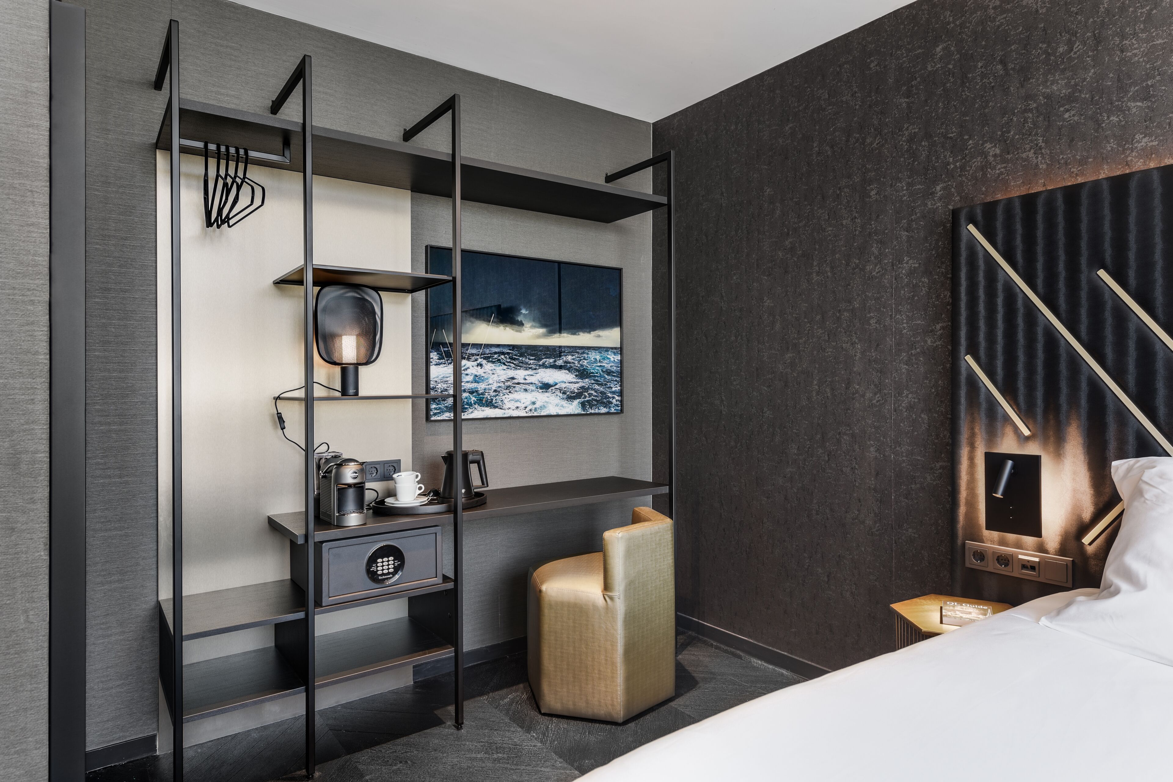 A modern hotel room with a bed, a desk with a chair, a TV, and a wall-mounted shelf with a picture.