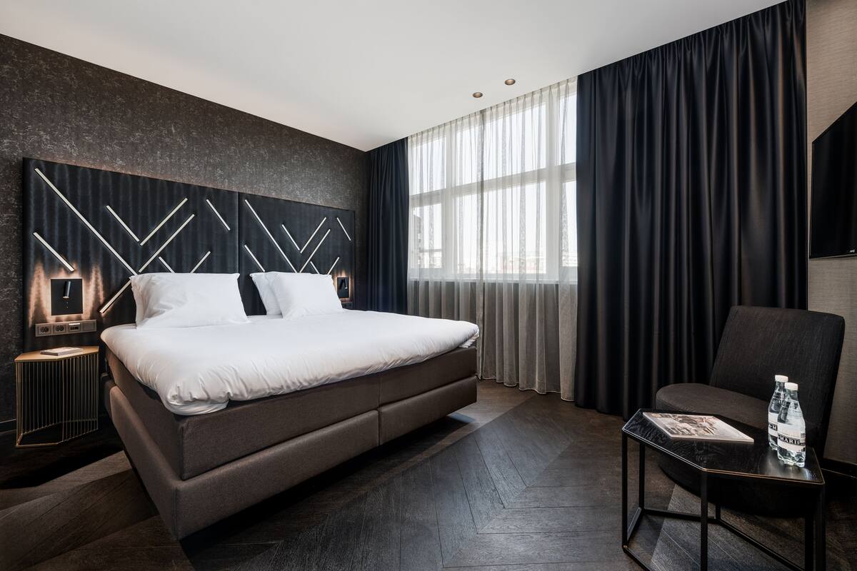 Ein modernes Hotelzimmer mit einem großen Bett, einem Nachttisch, einem Sessel und einem großen Fenster mit Vorhängen.
