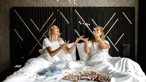 Cama com roupa de cama branca e objetos espalhados, incluindo uma revista e pipoca.