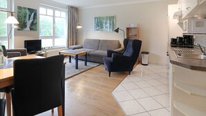 En moderne stue med sofa, lænestol og sofabord, et køkken med komfur og vask, samt store vinduer med gardiner.