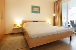 Een hotelkamer met een houten bed, twee bedlampen, een klein vloerkleed, een schilderij aan de muur en uitzicht buiten door een raam met gordijnen.