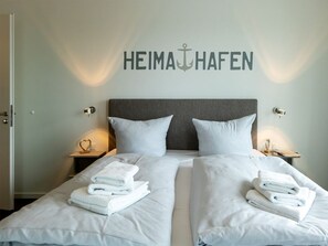 Ein Hotelzimmer mit zwei Betten, einem grauen Kopfteil, weißer Bettwäsche und einem an der Wand befestigten Schild mit der Aufschrift 'HEIMAHAFEN'.