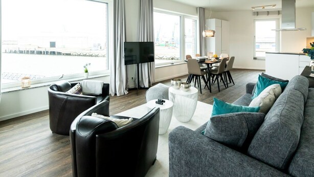 A modern living room with a grey sofa, black leather armchair, and a dining area with a table and chairs. - Hafenspitze App. 16 Pier 2.16 (Wismar)