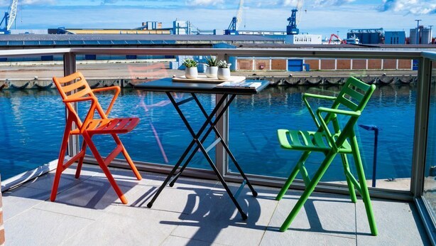 Un balcone con tavolo e due sedie che si affaccia su una distesa d'acqua con edifici industriali sullo sfondo.