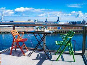 Un balcon avec une table et deux chaises, donnant sur une étendue d’eau, avec des infrastructures industrielles en arrière-plan.