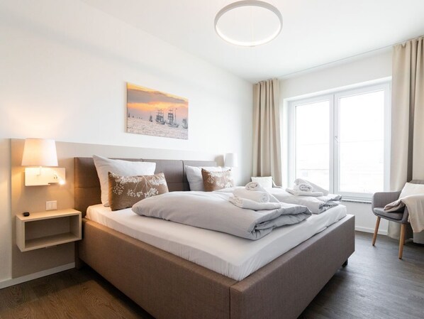 Une chambre d’hôtel moderne dotée d’un grand lit, de tables de chevet, d’une petite table, d’une chaise et d’une grande fenêtre avec des rideaux.