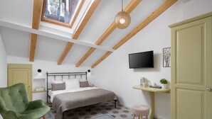Une chambre à coucher avec un lit, un fauteuil vert, une petite table, un téléviseur et une fenêtre ornée de poutres en bois.