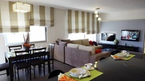 A modern living room with a sofa, dining table, and chairs. There is a television mounted on the wall and a window with curtains. - Eco Two bed Apartment With Roof Garden in Amman (Amman)