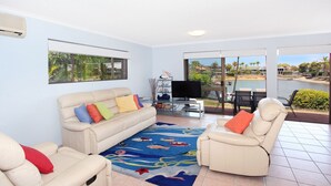 Un salón con un sofá beige, cojines coloridos, un televisor de pantalla plana y vistas a una playa a través de puertas de vidrio grandes.