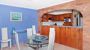 Un comedor con una mesa y sillas de vidrio, pared azul y cocina de ladrillo con armarios de madera.