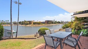 Una terraza con mesa y sillas que da a una masa de agua, palmeras y edificios al fondo.