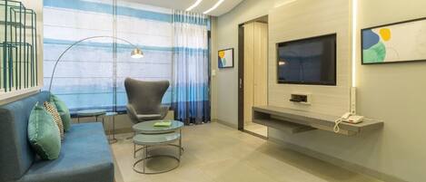 Una sala de estar moderna con sofá, sillón y mesa de centro, un televisor de pantalla plana montado en la pared, y un ventanal con cortinas.