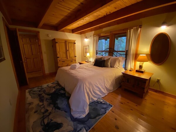 A bedroom with a large bed, wooden flooring, a nightstand, and a window with curtains.