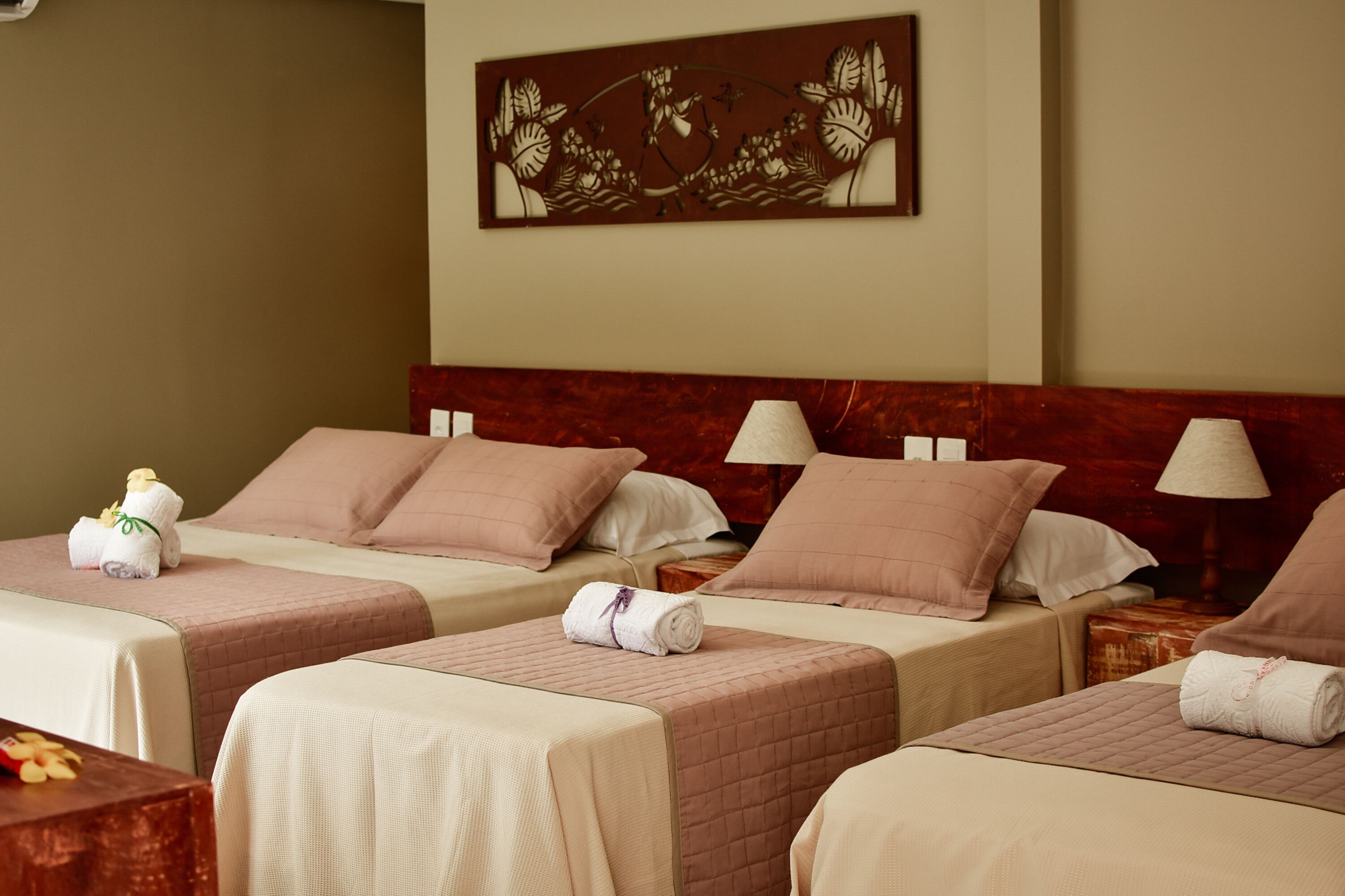A hotel room with three beds, each with a pink quilted bedspread and white pillows. There is a decorative wall piece above the beds and a small table with a lamp and a teddy bear on the left side.