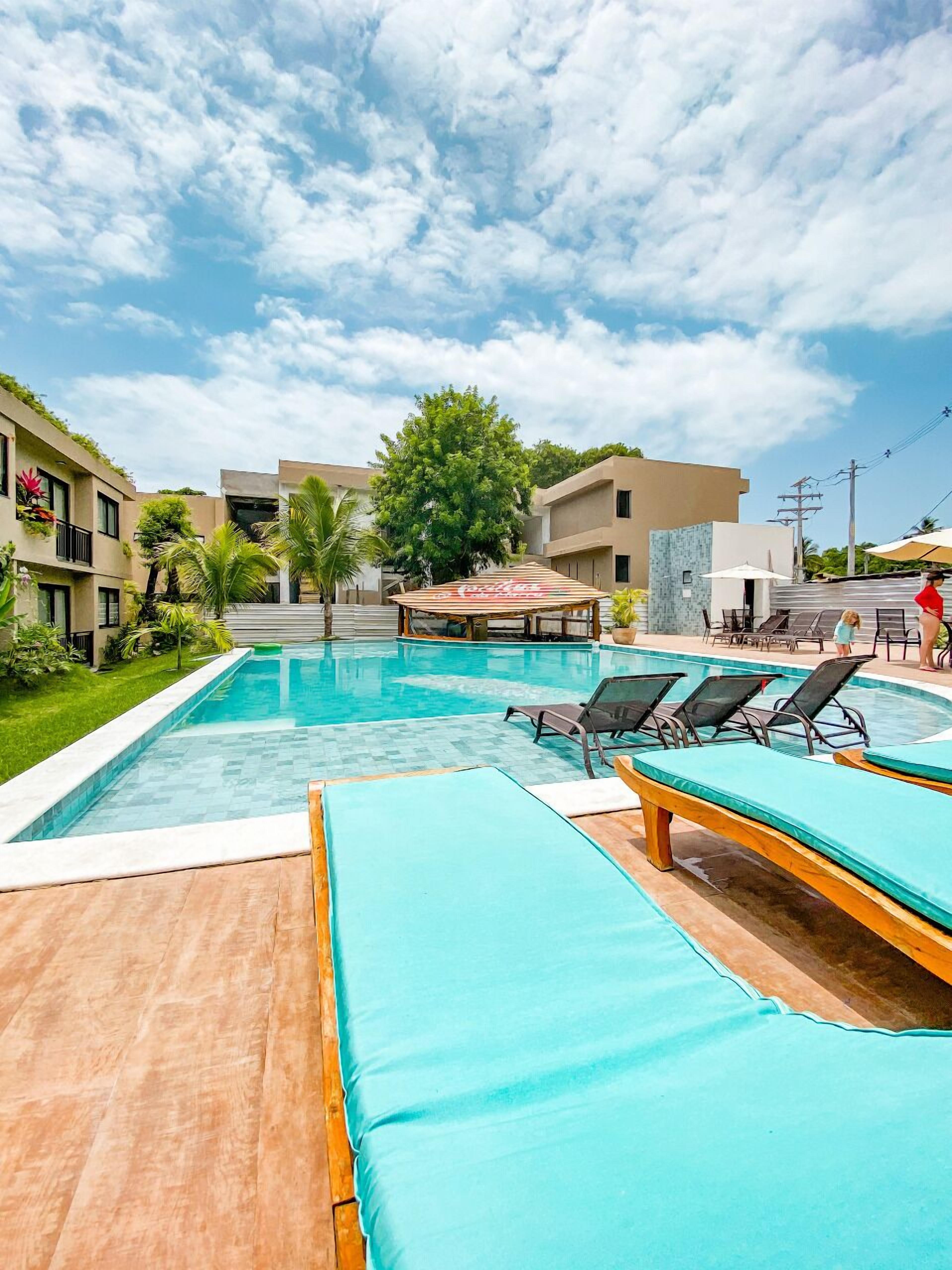 En swimmingpool med solstolar och ett avskärmat sittområde, omgiven av byggnader och grönska.