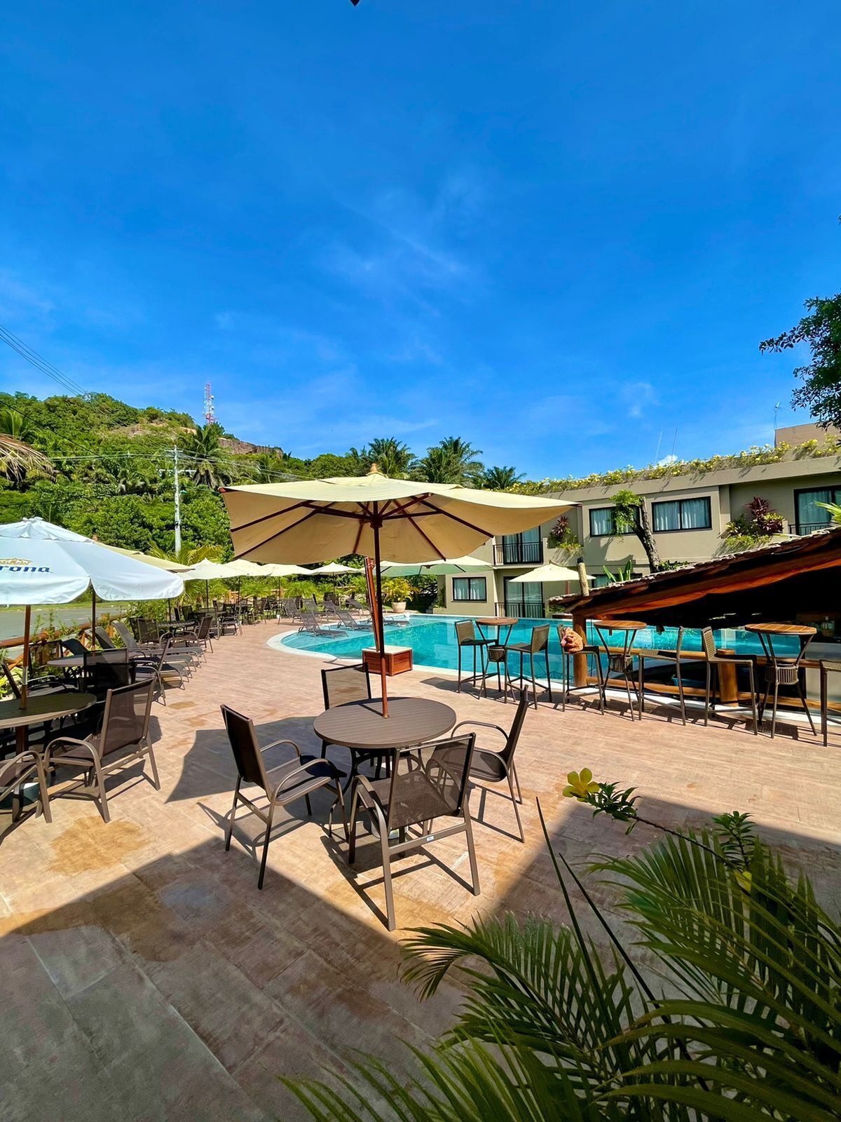 Uma área na beira da piscina com mobiliário de exterior e guarda-sóis, rodeada por vegetação e uma encosta.